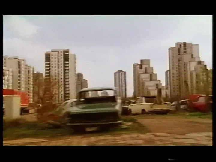A wrecked van and abandoned cars on waste ground below a skyline of concrete high-rise blocks