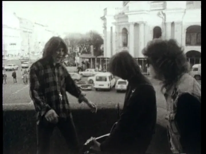 Three young people talking on a terrace above a busy street, a colonnaded church behind them