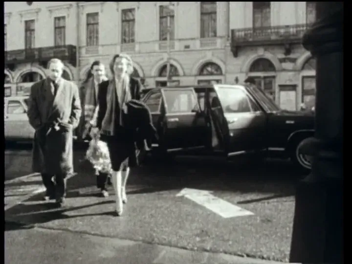 A delegation walking away from a black limousine outside a grand nineteenth-century facade, a woman in front carrying a bouquet
