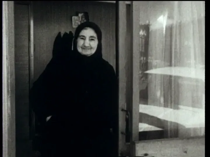 An elderly woman in a dark headscarf and coat standing smiling in the open doorway of her flat